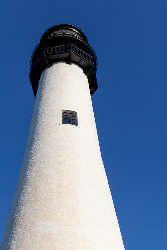 A tall, white Cape Florida Lighthouse by FN Prints stands against a clear blue sky, featuring a single window and a black observation deck at the top. This color photography print captures an upward view of the structure, emphasizing its height and slender design on Key Biscayne.