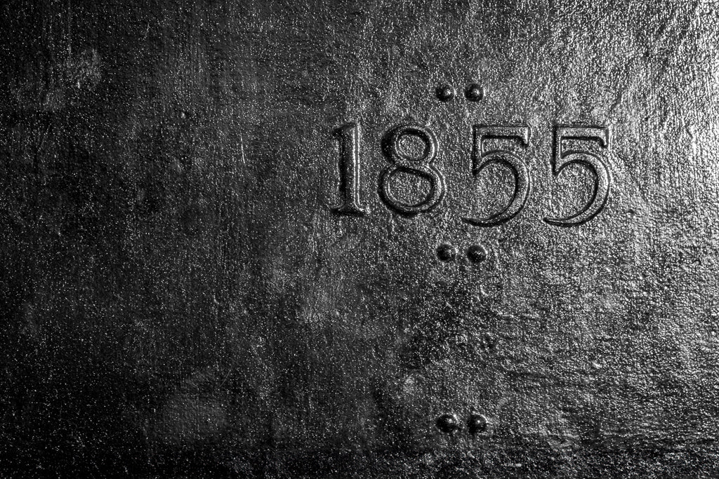 The 1855 by FN Prints showcases a dark, textured surface with embossed metal numbers "1855" at its center. Vertical arrangements of four small dots in two sets flank either side, enhancing the rough, metallic aesthetic. This antique industrial design makes it ideal as an abstract photography print or a striking piece of industrial wall art.