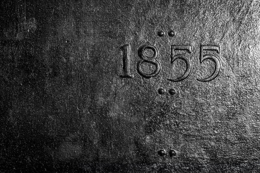 The 1855 by FN Prints showcases a dark, textured surface with embossed metal numbers "1855" at its center. Vertical arrangements of four small dots in two sets flank either side, enhancing the rough, metallic aesthetic. This antique industrial design makes it ideal as an abstract photography print or a striking piece of industrial wall art.