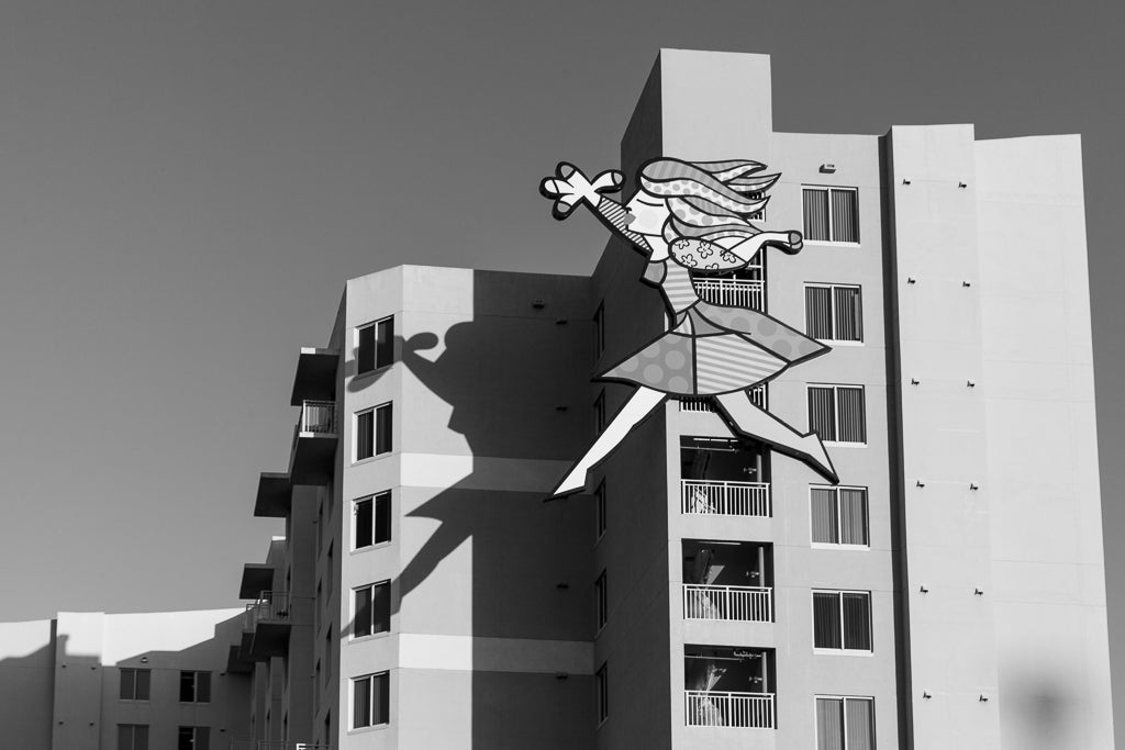 The black and white photo captures a tall building with balconies and windows. Attached to the building is an art installation called "Prancing Lady" by FN Prints, featuring a large, colorful cartoon-like figure of a woman holding a flower, partially casting a shadow on the building's surface.