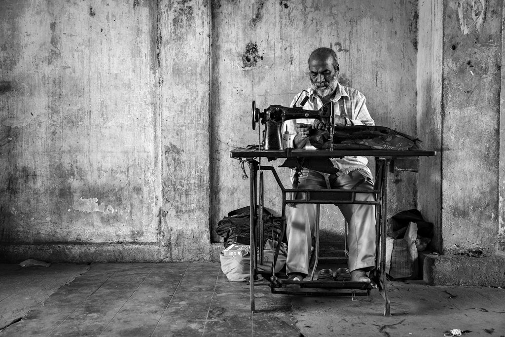 A black and white image from FN Prints' Street Seamster: Black & White series depicts an elderly man seated at a vintage sewing machine, deeply focused on his work. The backdrop is a worn and textured wall, surrounded by various bags and cloth items. This scene beautifully captures the essence of Goan craftsmanship and artisanal workmanship.