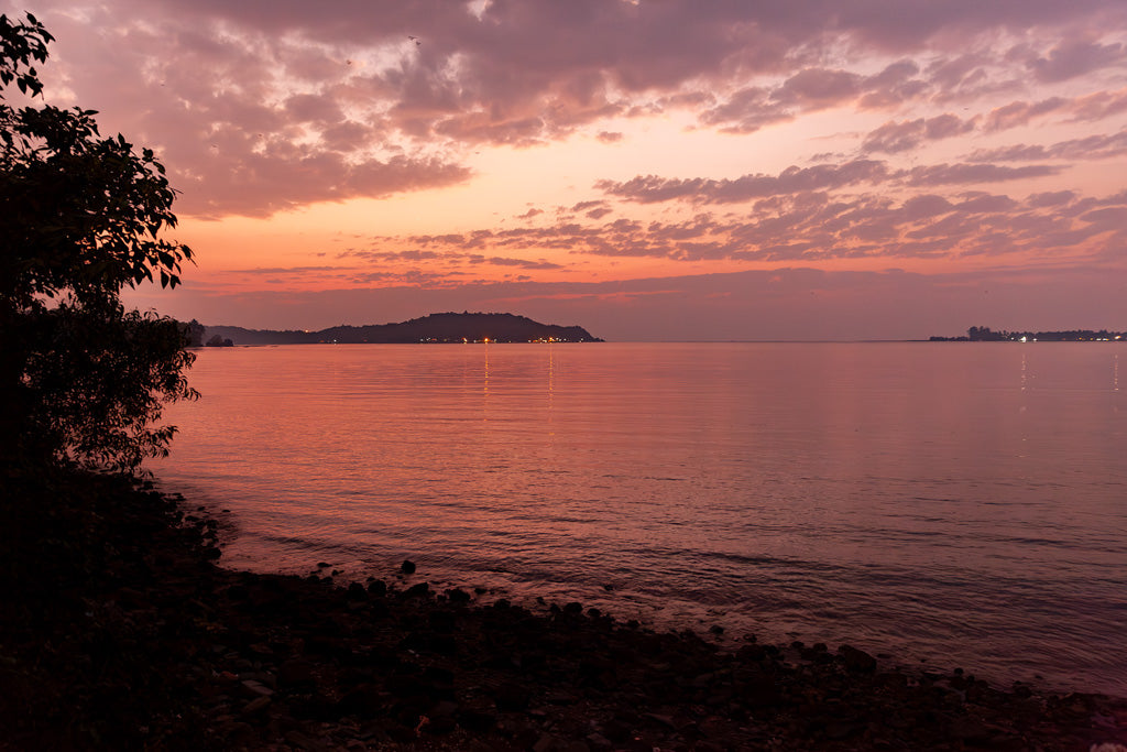 The "Rose Gold Sunset" print by FN Prints beautifully captures a tranquil scene on a Goan beach, featuring a calm body of water mirroring the sky painted in shades of pink, purple, and orange. Silhouettes of trees frame the left side of the image, while distant islands are visible on the horizon.