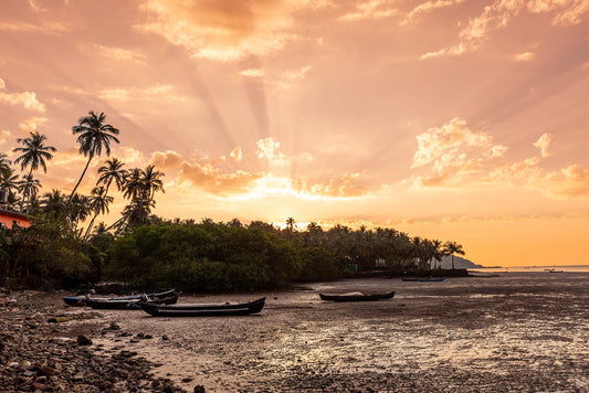 The "Goan Beach Sunset" by FN Prints features a tranquil tropical sunset with golden-orange skies. Sun rays gently filter through the clouds, casting a warm glow on palm trees and a shoreline dotted with moored boats. This photograph beautifully captures the calm reflection of the warm sky on the water, creating a serene and picturesque view brimming with coastal charm.