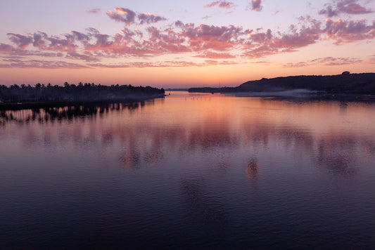 The FN Prints "Chapora River Sunset" beautifully captures a serene evening waterscape, where the tranquil Chapora River mirrors the pink and purple hues of the sunset. The sky is embellished with scattered clouds, while trees and hills silhouette the horizon in the distance. A gentle mist envelops parts of the shoreline, enhancing its twilight charm.