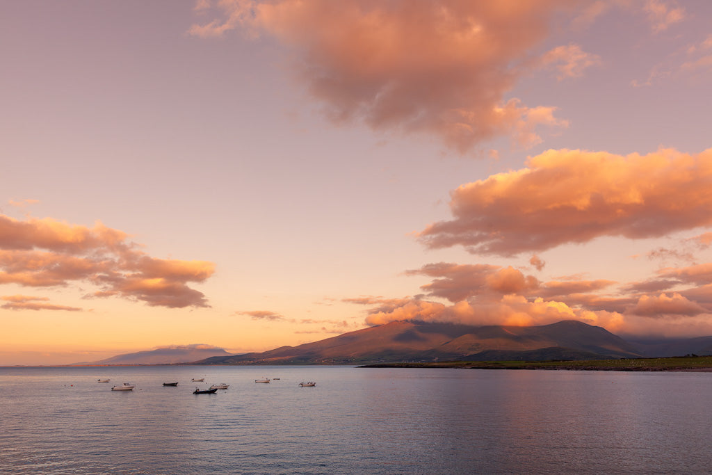 A tranquil coastal scene featuring the "Brandon Bay" at sunset with a few small boats anchored in calm waters. The sky is painted in hues of orange and pink, with scattered clouds. In the background, rolling hills and mountains are partially covered in soft clouds, creating a perfect setting for Ireland sunset photography captured by FN Prints.