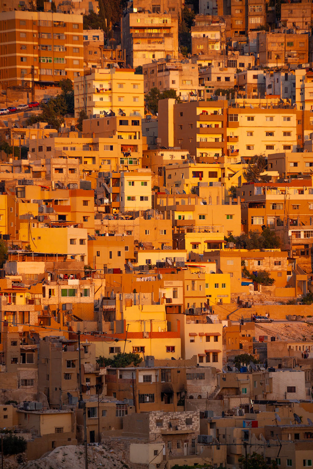 Title: Jabal Amman Sunset

Description: The FN Prints captures a densely packed hillside cityscape featuring an array of rectangular buildings painted in shades of yellow, orange, and beige. As the warm hues of the Jabal Amman sunset embrace the scene, the image vividly portrays the texture and ambiance of an urban area boasting a distinct Middle Eastern allure and architectural style.