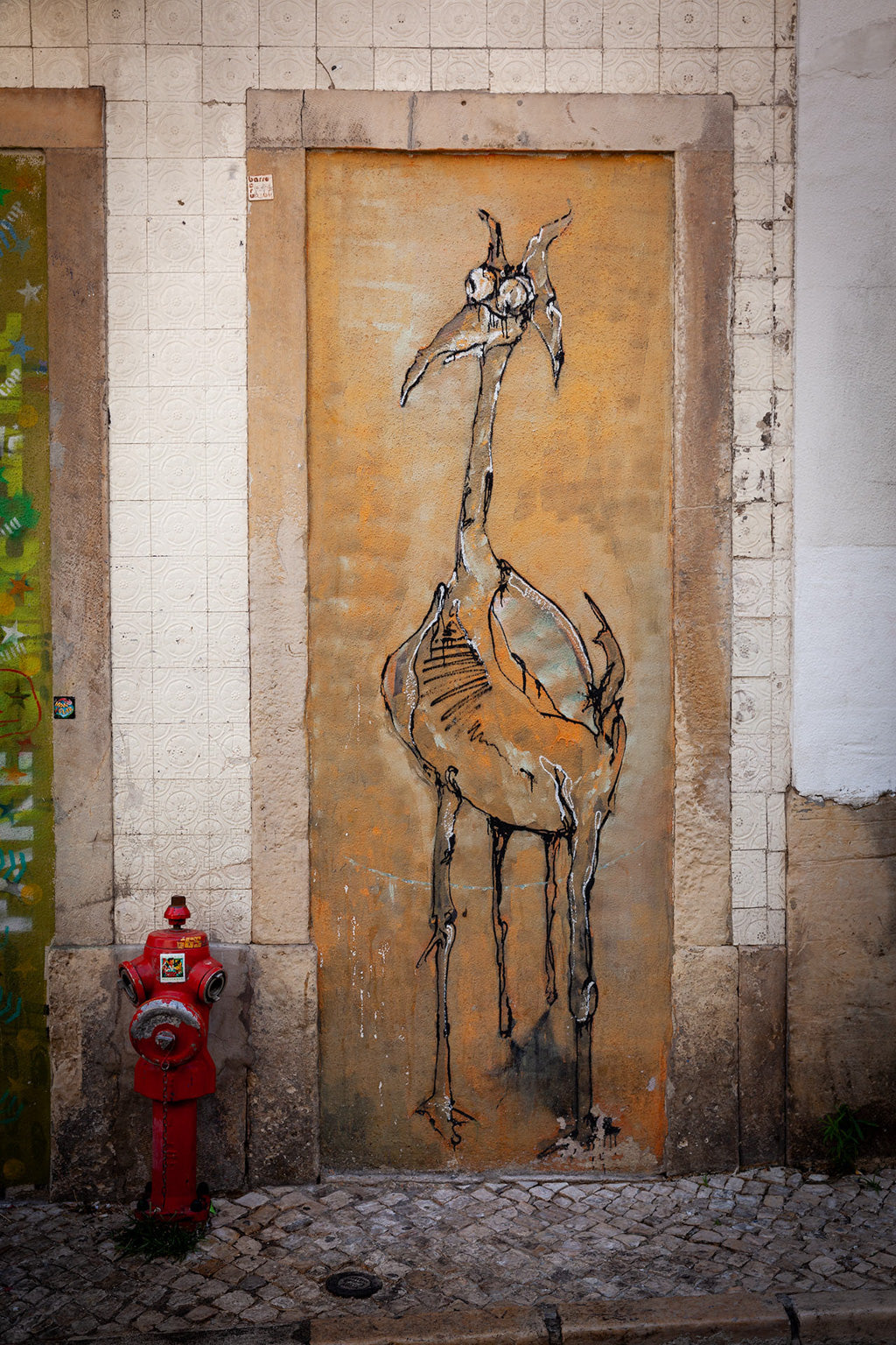 Lisbon Graffiti by FN Prints features a tall, abstract bird with long legs and a slender neck. This urban street art masterpiece is sketched in black lines against an orange-brown background. A red fire hydrant is situated on the cobblestone sidewalk in front of the artwork.