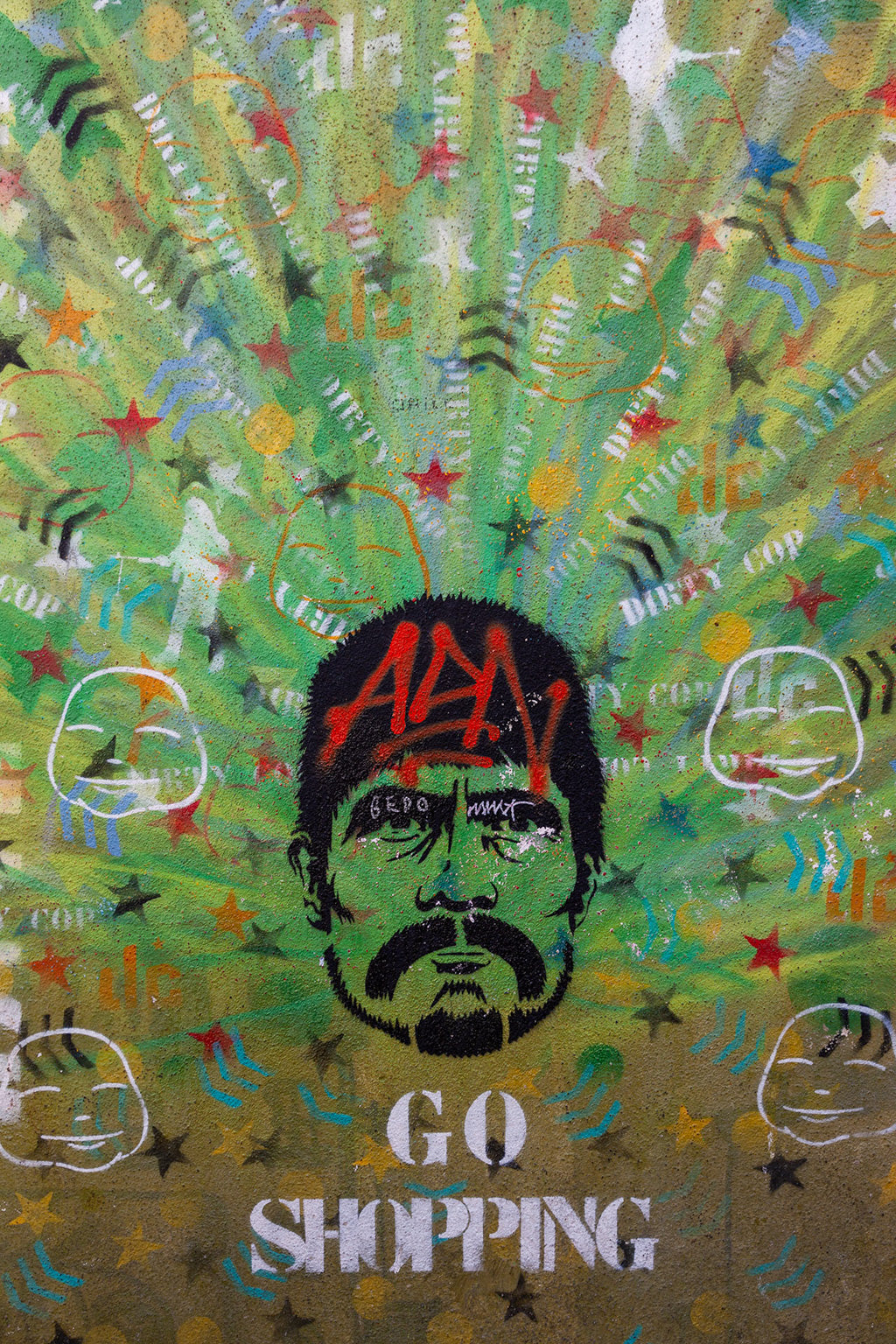 A vibrant showcase of urban creativity, Dirty Cop Colour by FN Prints centers on a man's face. Red spray paint partially conceals his features with "ADD" prominently written on his forehead. The background fuses assorted texts and images, highlighted by the stenciled phrase "GO SHOPPING" at the bottom.