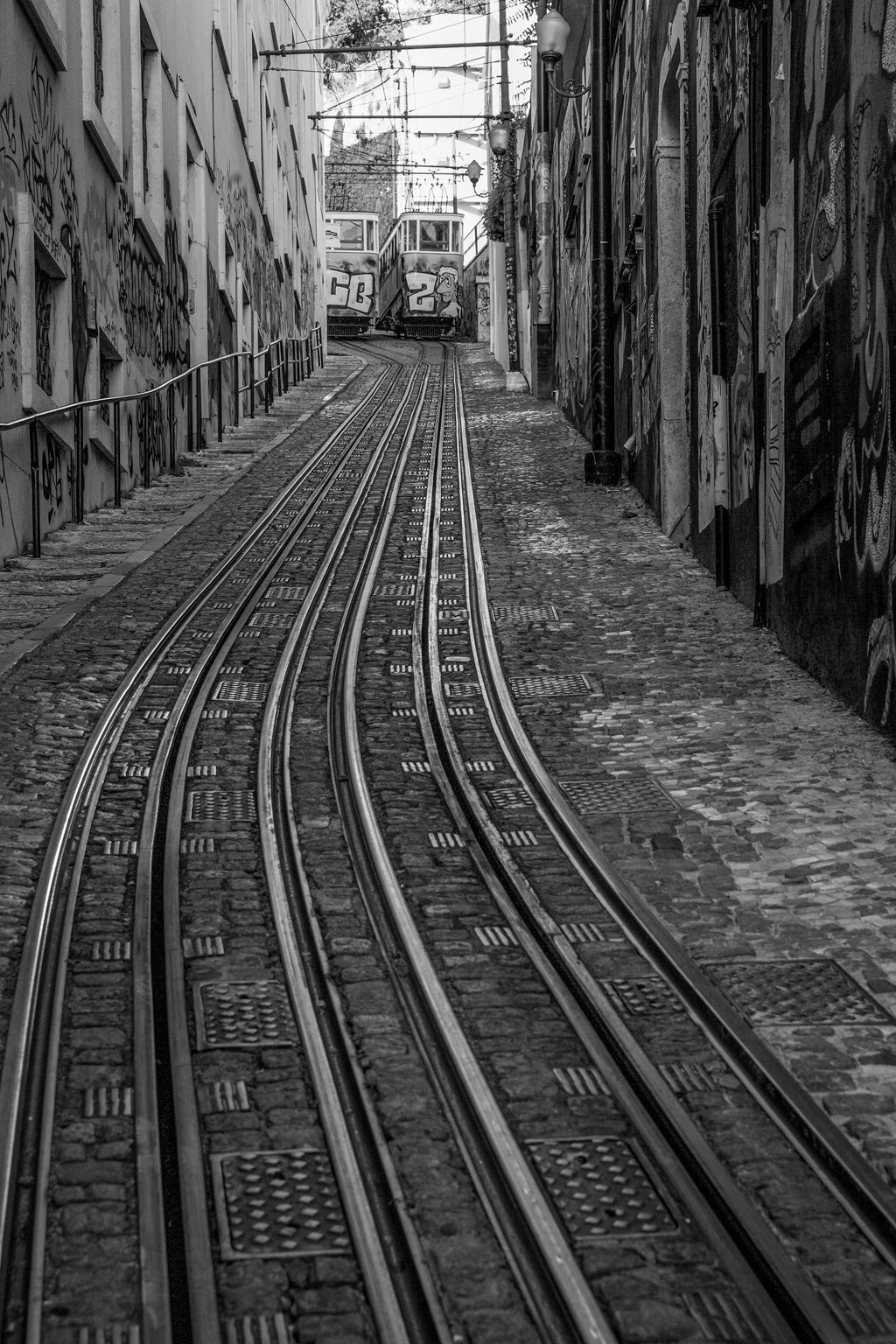 The FN Prints "Tram Tracks Black & White" features a black-and-white photograph of a steep, cobblestone street with tram tracks running through the center. Two graffiti-covered trams ascend the hill, flanked by buildings adorned with artistic graffiti on their walls.