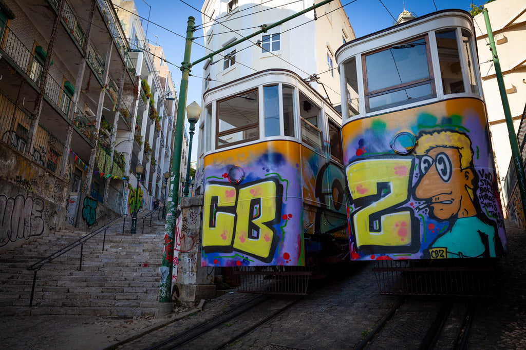 Two decommissioned trams, branded with FN Prints' "Decommissioned Trams Colour," pass each other on parallel tracks in a narrow, steep street lined with steps on one side. Old multi-story buildings adorned with additional graffiti art frame the scene. The vibrant urban artwork adds a splash of color to the Lisbon cityscape, transforming it into an engaging subject for urban photography.