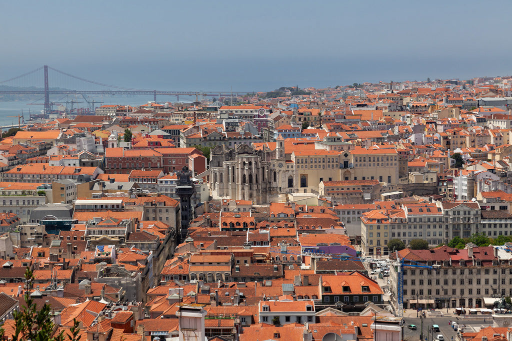 Aerial view of a historic Lisbon cityscape in "Lisbon Cityscape Colour" by FN Prints, featuring clustered orange rooftops and several notable buildings. The prominent 25th of April Bridge spans across the water in the background under a clear, blue sky.