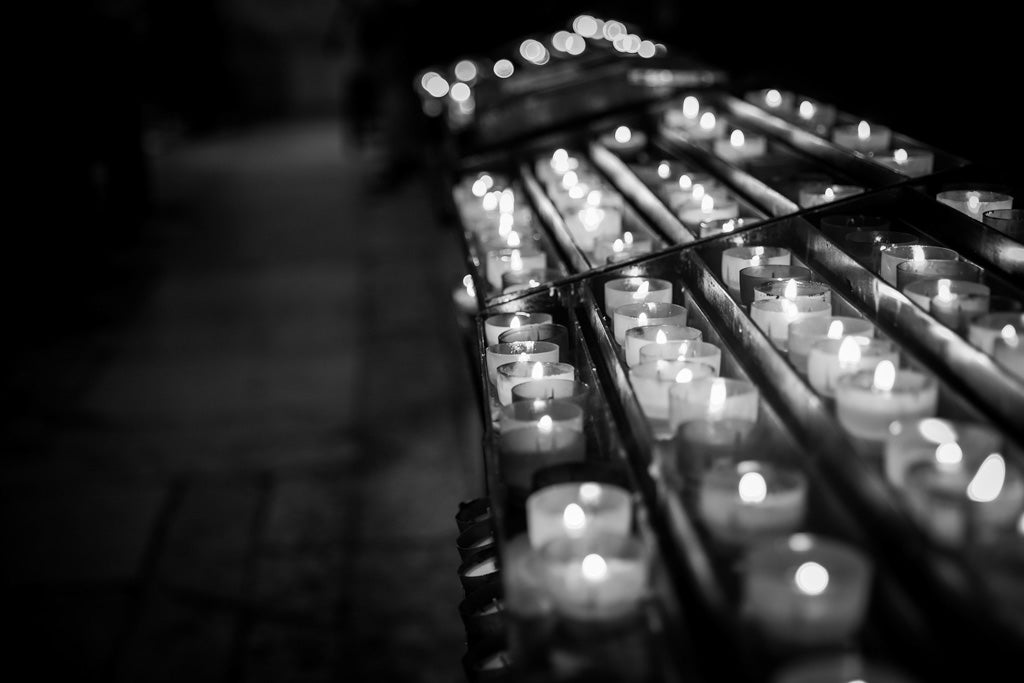 A black-and-white photograph captures FN Prints' Prayer Candles arranged in rows, each casting a soft glow. The candles rest on a metal rack and extend into the distance, enhancing the sense of depth. The dimly lit and blurred background emphasizes a calm and reflective atmosphere, reminiscent of Lisbon Cathedral.