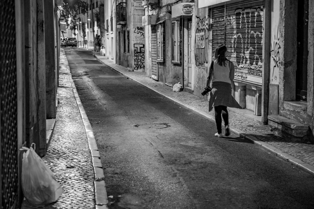 The FN Prints "Lisbon Photographer" is a black and white photograph that captures the essence of Lisbon's narrow urban streets at night. The scene features a person on the right side walking away with a camera, framed by graffiti, a closed shutter door, and scattered items like a plastic bag and barrel. This dimly lit setting perfectly evokes the spirit of urban exploration.