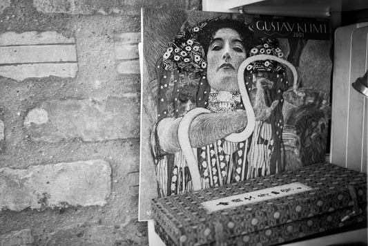 In a black and white abstract photograph, a shelf showcases the FN Prints product "Gustav Klimt & A Chinese Box," featuring a detailed drawing of a woman adorned with intricate patterns and jewelry. Below this captivating calendar, decorative boxes with geometric designs are neatly arranged. The wall adjacent to the shelf has an appealing texture with exposed bricks.