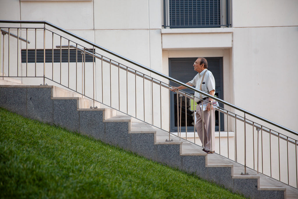 An older man wearing a light-colored shirt and pants climbs a concrete staircase bordered by a metal handrail, embodying urban elegance. Next to the stairs is a green grass slope, contrasting with the modern aesthetic of The Pause Colour white building with rectangular windows by FN Prints in the background.