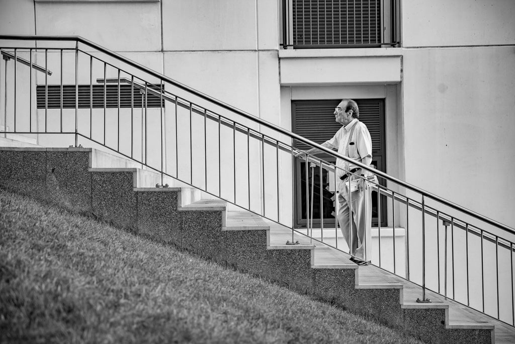 A man in a light-colored outfit walks up a flight of stone stairs with a metal railing. The background reveals the wall of a modernist building, complete with windows and vents. The photograph, exuding urban sophistication, is titled "The Pause Black & White" by FN Prints.