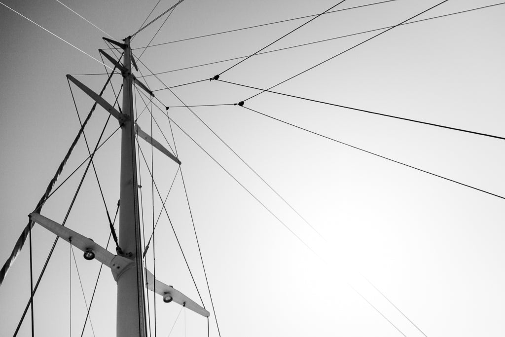 The Mast by FN Prints is a black-and-white photo focused on the mast of a sailboat. The image captures several lines and rigging extending from the tall mast against a clear, bright sky, highlighting the intricate network of ropes essential to its nautical design.