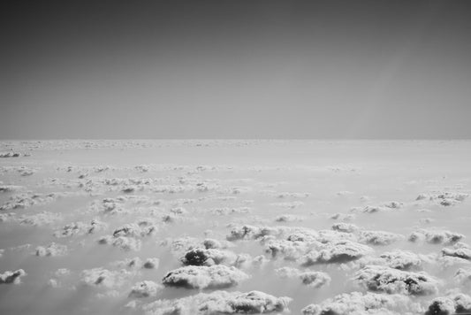 The FN Prints "Flying Above The Clouds" features a black-and-white aerial photograph that captures an expanse of fluffy clouds against a clear horizon. Taken from above, the cloud layer imparts a serene and endless feel to the sky, evoking a sense of calm and vastness through its monochromatic tones.