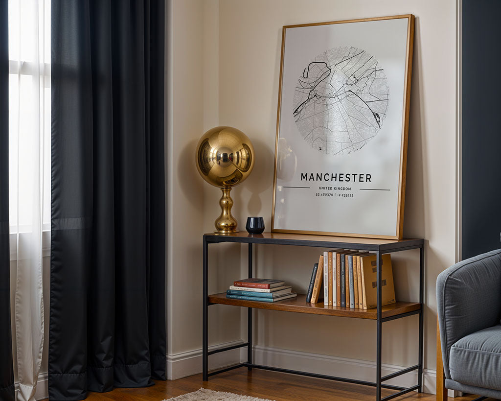 A FN Prints Manchester City Map hangs on the wall next to a gold lamp, resting on a two-tier console table with books and a black candle. Urban design features include sheer and dark curtains framing the window, casting soft shadows over part of a gray armchair.