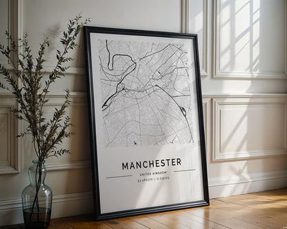 The FN Prints Manchester City Map, a framed black-and-white poster with coordinates, leans against a white paneled wall as an urban design art piece. Sunlight creates shadows on the wooden floor, while a nearby vase with branches brings nature into the scene.