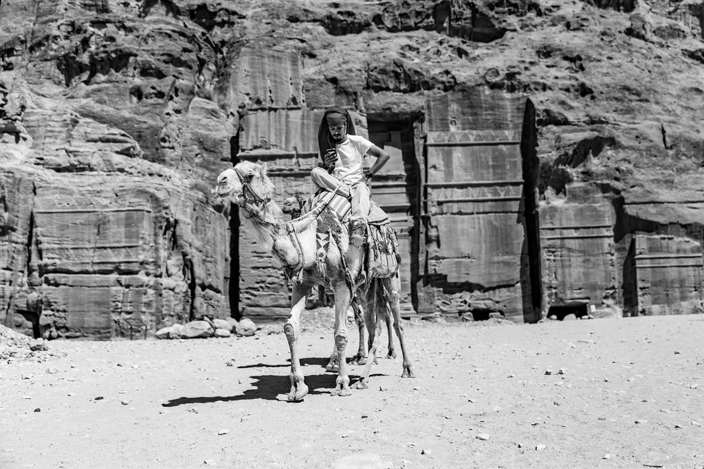 The Modern Bedouin Black & White print by FN Prints portrays a person riding a camel, possibly holding a mobile phone. The backdrop showcases stunning ancient rock formations and carved structures that evoke a historical site. The sandy, arid ground enhances the desert setting, highlighting the blend of traditional and modern elements.