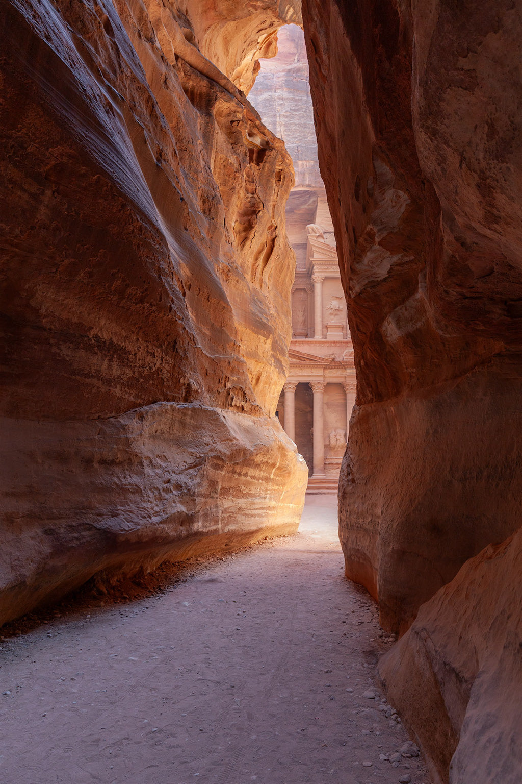 A Peak Of Al-Khazneh - The Treasury Petra Colour by FN Prints captures a narrow passageway between the tall, rocky canyon walls of Petra, Jordan. The sunlight illuminates the scene, revealing columns of the ancient Al-Khazneh (The Treasury) in the distance. The sandy pathway and light accentuate the rich red and orange hues of the stone.