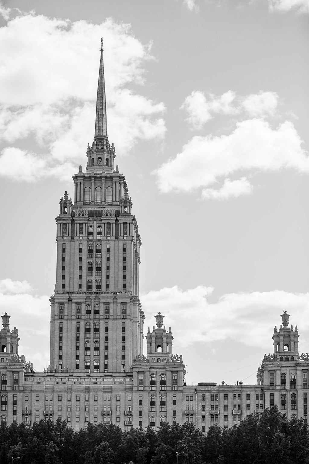 Black-and-white photograph showcasing the grandiose Hotel Ukraina, Moscow by FN Prints. This multi-story building features a central tower with an ornate spire, encapsulating neoclassical architecture. The iconic structure stands proudly against a partly cloudy sky, with trees lining the foreground and framing this quintessential piece of the Moscow skyline.