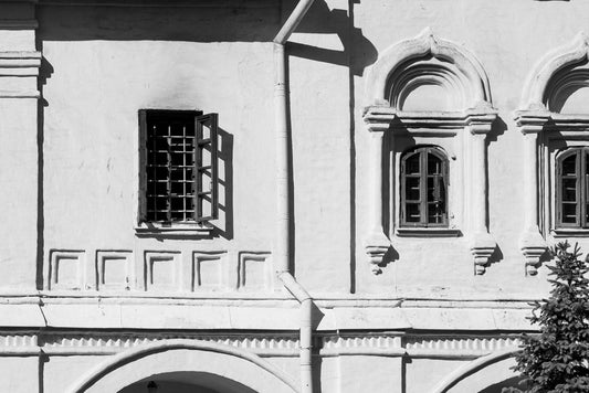 A monochrome photograph from FN Prints' "Russian Windows" collection captures the architectural elegance of an old Russian white stone building facade, adorned with arched windows and intricate decorative details. One window on the left is open with bars, while the two windows on the right are closed. A vertical drainpipe runs between the open and closed windows.