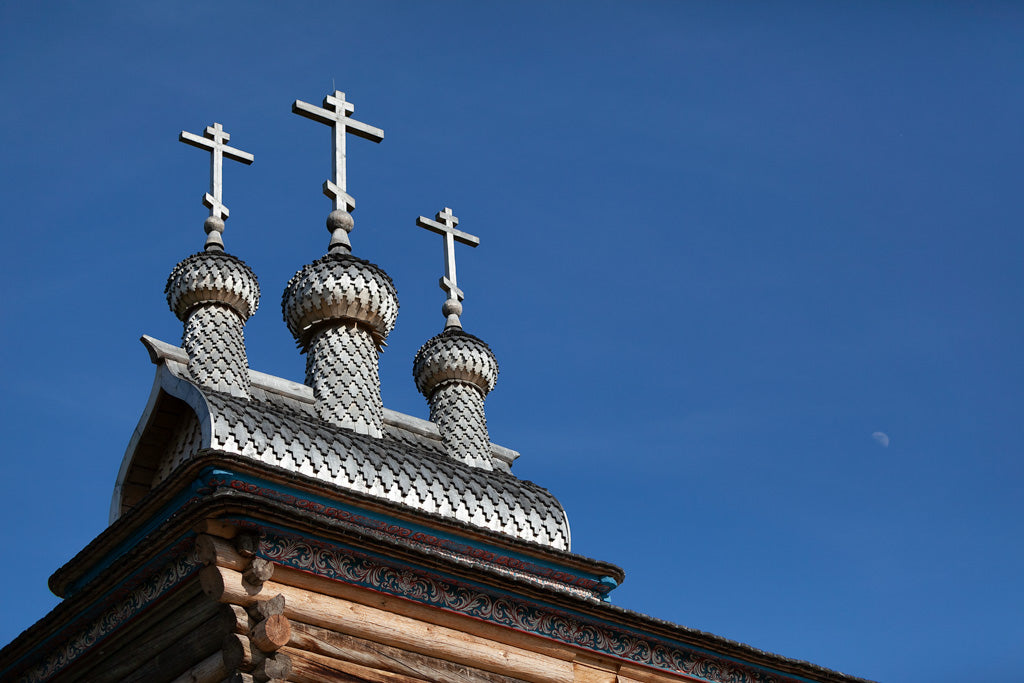 The image from FN Prints' "Three Crosses" shows the top part of a traditional Russian wooden church with intricate shingle patterns and three silver domes, each crowned with a wooden cross. This stunning example of religious architecture mirrors the elegance seen in places like Saint George in Moscow. The sky above is clear and blue, with a faint moon visible in the background.