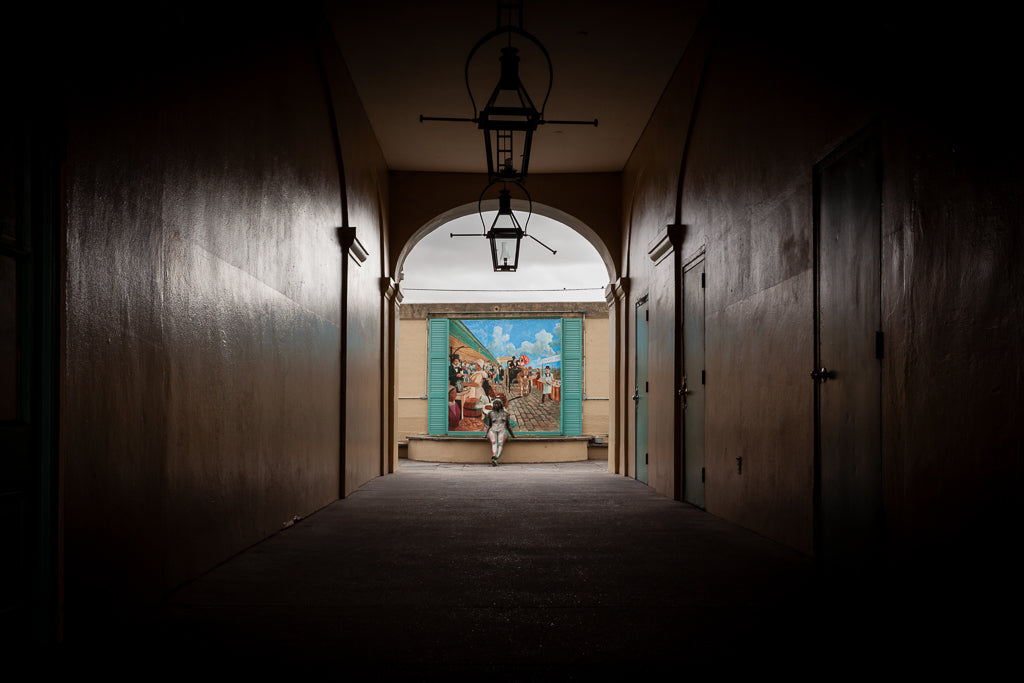 A person stands alone in a dimly lit hallway with beige walls, beneath two overhead lanterns. The hallway opens up to "Michelle And The Mural" by FN Prints, a vivid depiction of a French Market scene showcasing people against a blue sky with clouds. Pale green shutters frame this stunning mural at the end of the corridor.