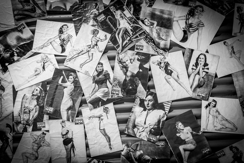 A scattered collection of black-and-white vintage pin-up photographs featuring women in various poses and outfits, some images worn and crumpled to enhance a nostalgic, aged feel. This evocative black-and-white photography print from FN Prints, titled "The Pin Up Girls: Nostalgia & Dreams," perfectly captures the essence of pin-up culture.