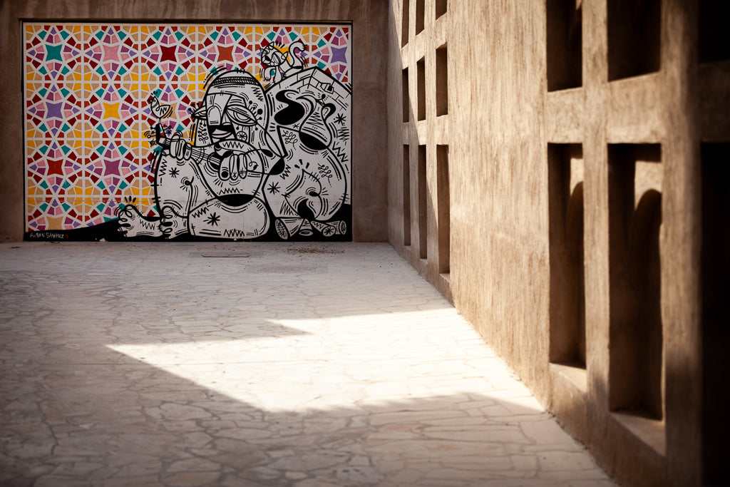 An intricately painted Rubén Sánchez mural featuring abstract human figures with patterns is displayed on a textured wall. The background consists of colorful geometric shapes in a mosaic style. The surrounding area boasts a shadowy, textured walkway and square cut-outs, reminiscent of FN Prints' "Bastakiya Wall, Dubai 2013.