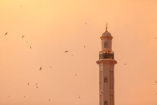 Minaret of Dira mosque in Dubai with birds flying around it at sunset. 