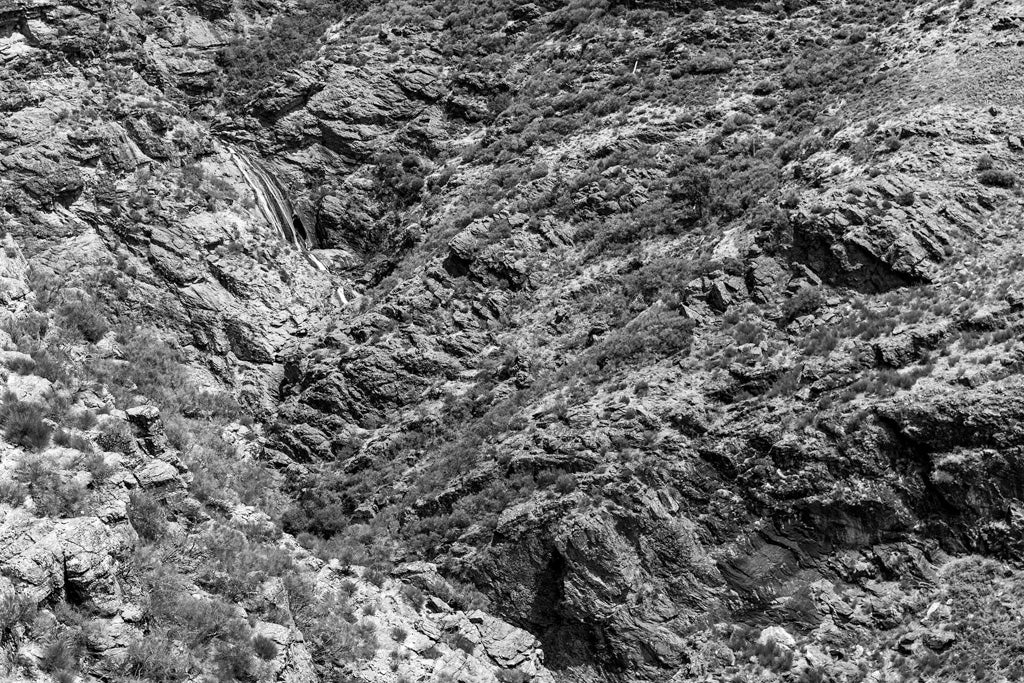 A black-and-white photograph from FN Prints, titled "Delicate Waterfall," captures the rugged landscape of Portuguese mountains, showcasing steep cliffs and sparse vegetation. The terrain appears dry and craggy, with various rock formations creating a textured and dramatic natural scene.