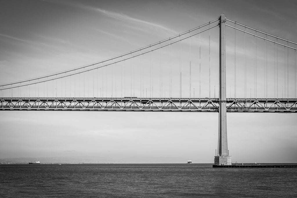 A black and white photograph from FN Prints, titled "Golden Gate Tower," beautifully captures the architectural elegance of a large suspension bridge reminiscent of the Golden Gate Bridge. The side view showcases its tall towers connected by numerous cables under a clear sky, with a distant ship partially visible on the horizon.