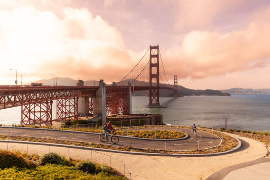 The Golden Gate Cyclist photography print by FN Prints captures the Golden Gate Bridge in San Francisco under a sky with gentle pink and orange clouds. In the foreground, cyclists ride along a winding path, with the iconic red bridge and the bay prominently displayed in the background.