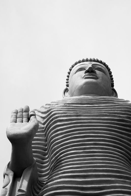 A low-angle black and white close-up photograph titled "Lord Buddha: Tsunami Honganji Viharaya" by FN Prints. The statue's right hand is raised with the palm facing outward, a gesture symbolizing teaching or fearlessness. The serene face is framed by a textured robe and curly hair.