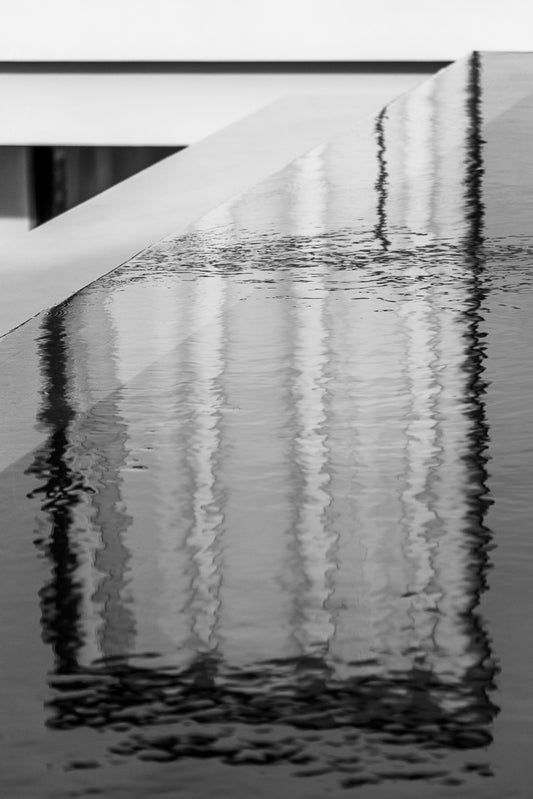 Abstract Reflections by FN Prints features a black-and-white photo of a reflective water surface on an angular structure. The reflections of vertical lines and contrasting geometric shapes create a striking, abstract image. This modern and minimalist scene makes the perfect wall art.