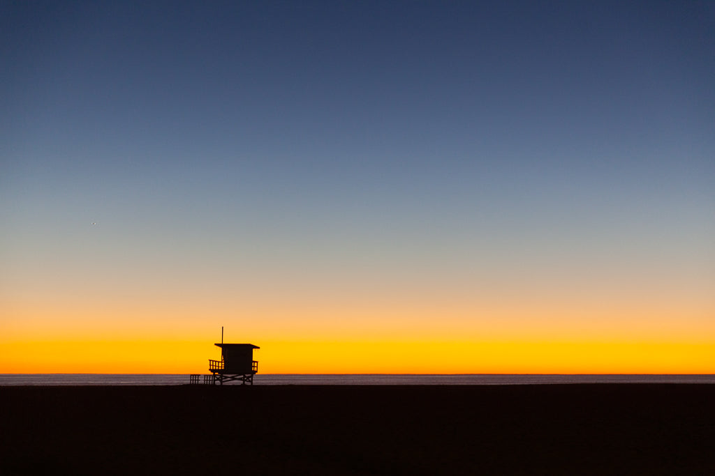 A captivating silhouette of "The Lifeguard Tower" by FN Prints adorns a Los Angeles beach against a vibrant sunset. The sky transitions from deep blue at the top to a bright, golden yellow near the horizon, casting reflections on the calm ocean below. The darkened beach enhances the vivid hues of the sky—making it perfect coastal wall art.