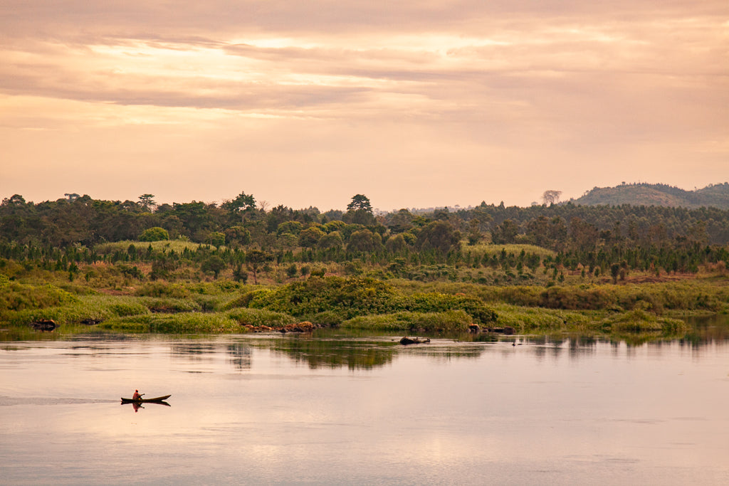 The FN Prints "Ugandan Canoeist" photography print beautifully captures a canoeist rowing on a calm Ugandan river during the golden hour sunset, with verdant trees and shrubs adorning distant hills under a softly lit sky with scattered clouds.