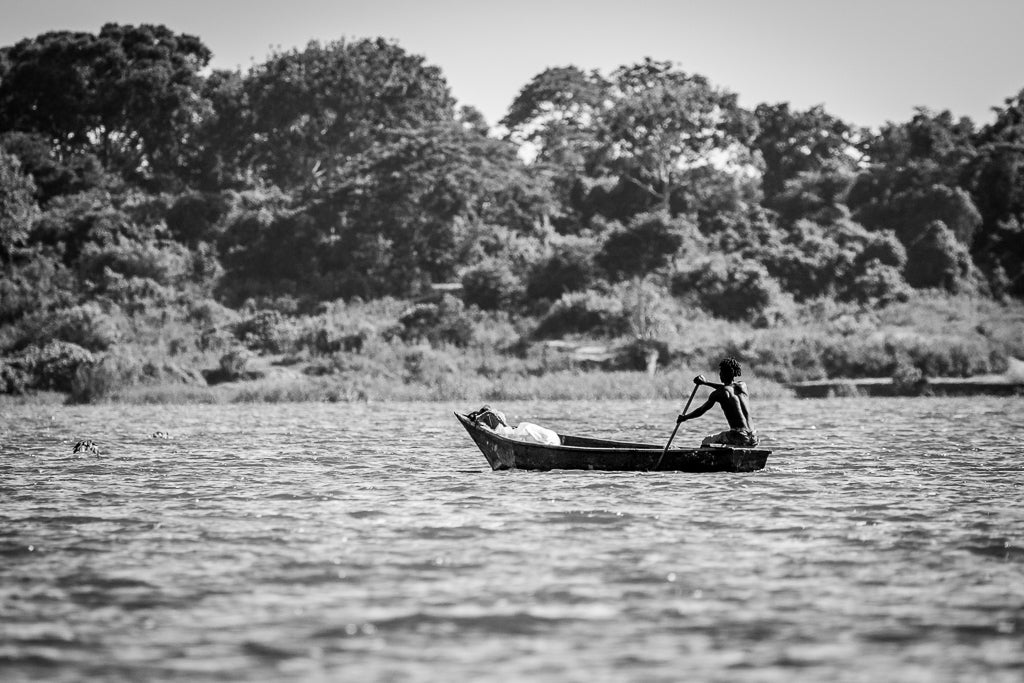 In FN Prints' "Jungle's Edge" black and white photography print, a person paddles a small wooden boat on a vast body of water. The background showcases dense, leafy trees and vegetation along the shoreline. The water appears slightly rippled, and the sky is clear. This piece makes perfect wall art for your home decor.