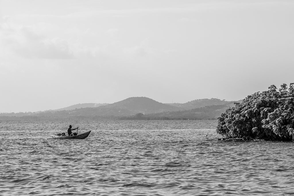 FN Prints' 'Lake Victoria Canoeist' captures a black and white image of a canoeist rowing a small boat on Lake Victoria. Hills can be seen in the distance, with a large shrub or tree on the right side. The horizon is hazy, perfectly conveying the serene and calm essence typical of fine art photography.