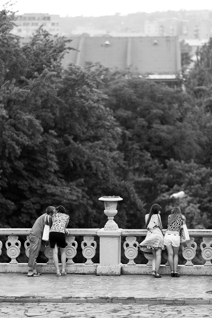 The FN Prints "Lovers and Friends" artwork captures a black and white scene of two pairs of people leaning over a stone railing, gazing out at a view. Behind them, a dense cluster of trees ascends to city buildings in the background. Embracing urban living, the casually dressed individuals appear engaged in conversation, enhancing the scene's authenticity.