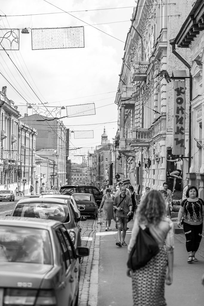 The Ukrainian Street print by FN Prints captures a black and white European scene with pedestrians walking along the sidewalk and several cars parked on the side. The street is lined with historical buildings, one of which features a vertical sign in Cyrillic script, epitomizing urban realism wall art in Ukrainian street photography.