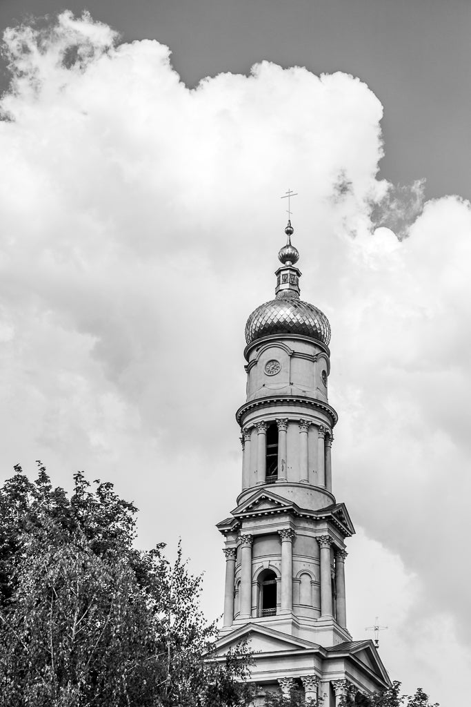 Black and white image of the Dormition Cathedral: Kharkiv's tall, ornate tower with a domed top adorned with a cross. The Neoclassical architecture features a clock and multiple arched windows. Leafy trees are visible in the foreground, and large, billowing clouds fill the background sky. This stunning artwork is offered by FN Prints.