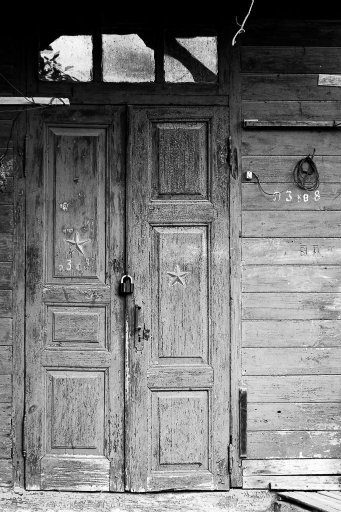 The weathered wooden door, known as "Locked Memories" by FN Prints, features two panels with stars in the center and is secured with a padlock. Exuding rustic charm, it forms part of an old structure with worn wooden planks and a small transom window above. To the right, a coiled wire and faded markings add to its vintage appeal—perfect for black and white photography.