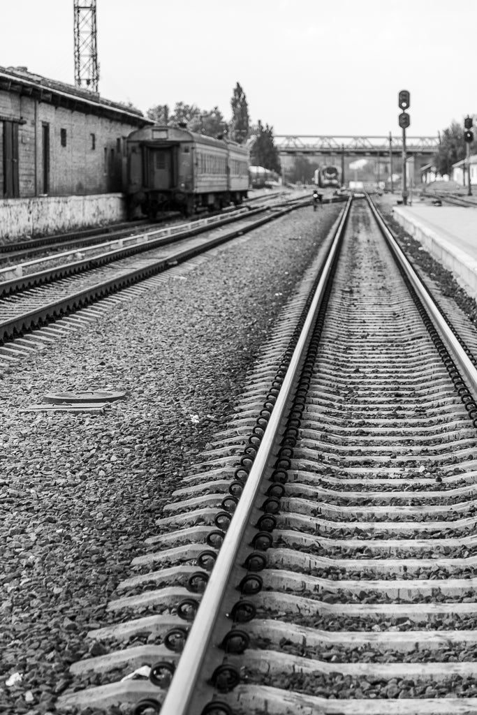 On the Tracks" by FN Prints captures train tracks leading to a distant station in black-and-white photography. The tracks run straight through a gravel-filled area with scattered debris. A train is parked on the left side near an old building, making this vintage wall art print timeless and evocative.