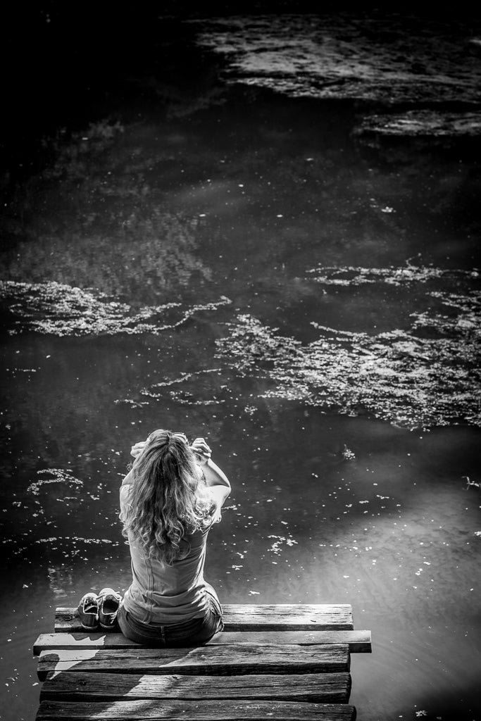 FN Prints' "Contemplative Waters" captures a serene moment in black and white, featuring a curly-haired individual seated on the edge of a wooden dock by a peaceful body of water. With their shoes placed beside them, the person seems to be taking a picture or looking through binoculars, embodying tranquil reflection. This piece is perfect for wall art.