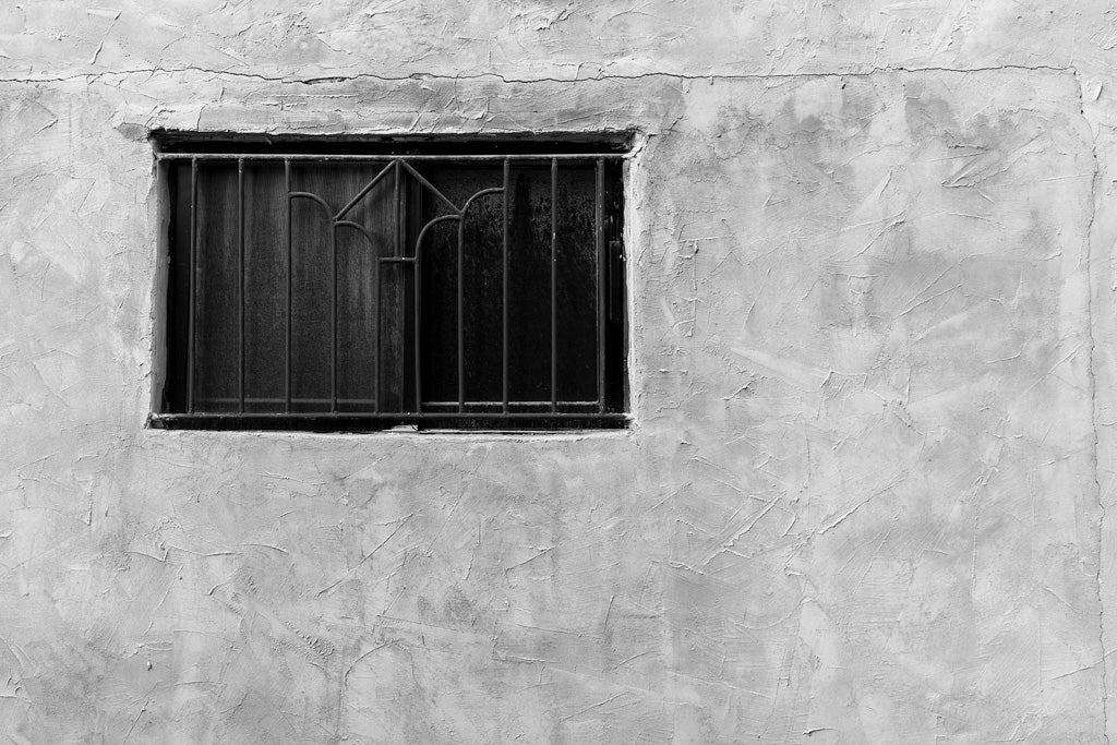 The Closed Window" by FN Prints features a small, rectangular window with metal bars embedded in a textured stucco wall, exuding urban sophistication. The window's bars arch gracefully at the top, while the weathered wall showcases visible cracks and varying textures. This black-and-white image captures timeless elegance amidst urban decay.