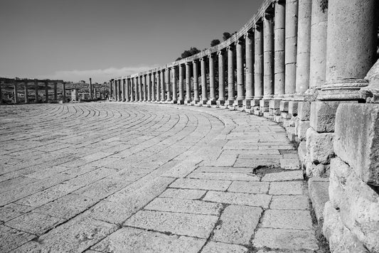 The FN Prints "Oval Plaza Jerash Black & White" captures a stunning black and white photograph of the large curved colonnade with ancient stone columns and a wide cobblestone path. The historic columns and pathway of the Oval Plaza in Jerash, Jordan extend into the distance, bordered by a low stone wall on one side.